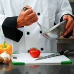 Caucasian chef mixing something in a bowl with fresh vegetables on a cutting board in front of him in a restaurant kitchen. Chef with vegetables